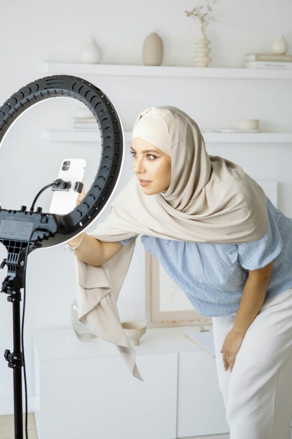 Stylish woman in hijab recording video at home with ring light and smartphone.
