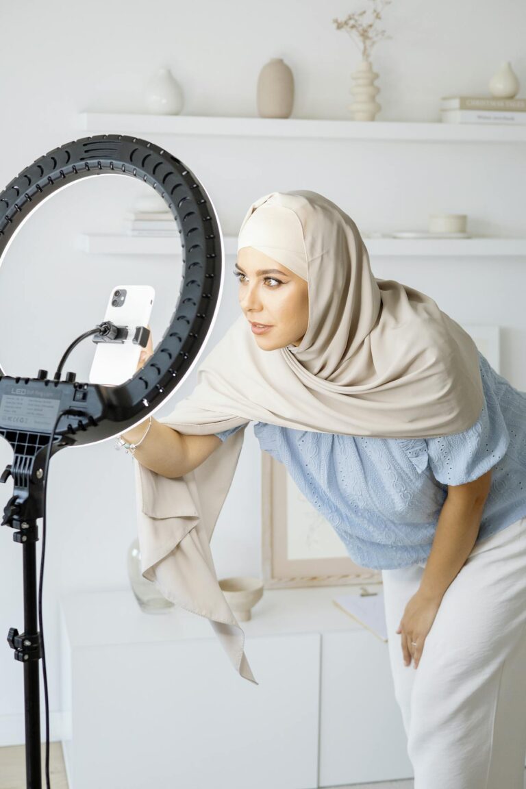 Stylish woman in hijab recording video at home with ring light and smartphone.